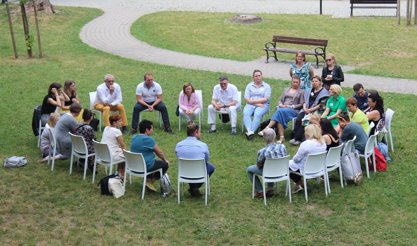 Erwachsene sitzen im Kreis auf Stühlen bei einem Treffen im Freien.